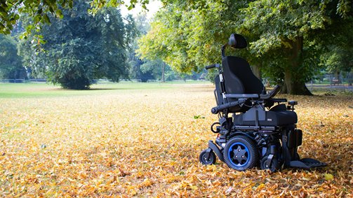 A mid wheel drive powered wheelchair in a park with floor covered with leaves.