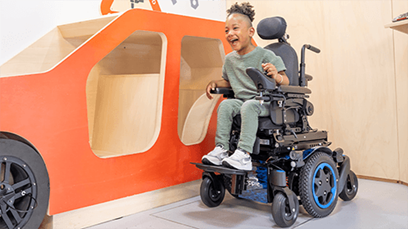 A young child sitting in a paediatric powered wheelchair with a friend next to them.
